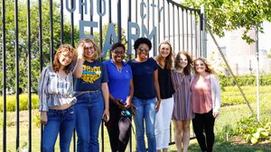 Seven young women stand smiling together in front of a metal gate that reads Ohio City Farm on a sunny day.