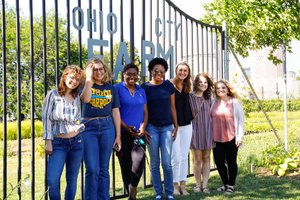 Seven young women stand smiling together in front of a metal gate that reads Ohio City Farm on a sunny day.