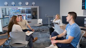 Four people sit in a computer lab, engaged in conversation with laptops and monitors around them.