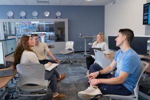 Four people sit in a computer lab, engaged in conversation with laptops and monitors around them.