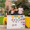 Four students stand behind a colorful table display with flags and photos on a college campus lawn.