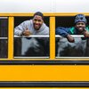 Two young men lean out of a yellow school bus window smiling and giving thumbs up.