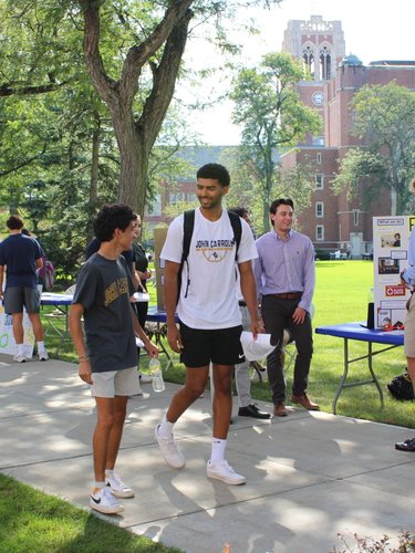 Students walk and talk along a shaded campus sidewalk lined with tables and posters on a sunny day.