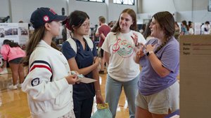 Four young women stand in a gymnasium talking, with one gesturing and others listening attentively.