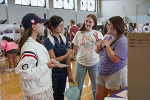 Four young women stand in a gymnasium talking, with one gesturing and others listening attentively.