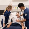 Two medical students in blue scrubs attend to a patient lying in a hospital bed with oxygen mask and monitoring devices.