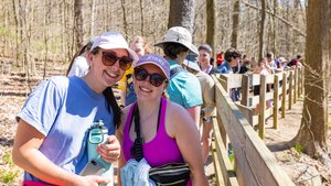 Two smiling women wearing sunglasses and hats stand near a wooden fence in a wooded area with a group behind them.