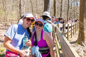 Two smiling women wearing sunglasses and hats stand near a wooden fence in a wooded area with a group behind them.
