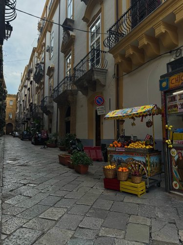 A narrow cobblestone street lined with tall buildings and a colorful fruit stand displaying oranges and lemons.