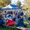 People gather and socialize under and around a blue tent labeled JCUSPORTS.COM and Blue Streaks on a sunny day.
