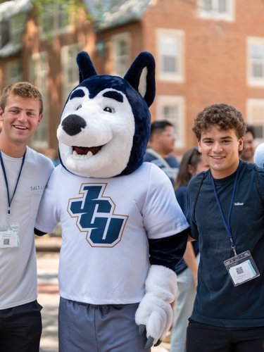 Two young men smile and pose with a person in a husky mascot costume wearing a JCU shirt outdoors.