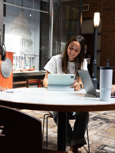 Three students sit around a round table working on a tablet, laptop, and notebook in a room with a large Capitol building.