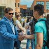 A man in a blue blazer and sunglasses shakes hands with a young man wearing a turquoise shirt and backpack outdoors.