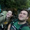 Two young people wearing backpacks smile at the camera while standing on a wooden bridge in a lush forest.