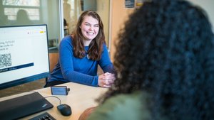 A woman in a blue sweater smiles warmly across a desk at another person with curly hair in an office setting.