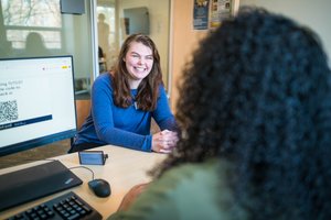 A woman in a blue sweater smiles warmly across a desk at another person with curly hair in an office setting.