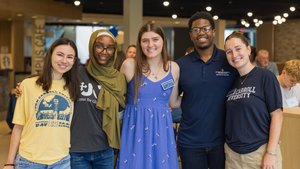 Five young adults stand close together smiling inside a well-lit room with other people in the background.