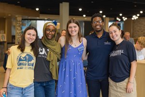 Five young adults stand close together smiling inside a well-lit room with other people in the background.