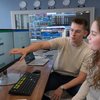A young man points at a computer screen while a young woman watches attentively in a room with multiple monitors and clocks.