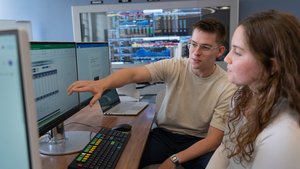 A young man points at a computer screen while a young woman watches attentively in a room with multiple monitors and clocks.