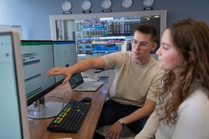 A young man points at a computer screen while a young woman watches attentively in a room with multiple monitors and clocks.