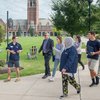 A group of six people walk and talk on a campus sidewalk near a green lawn and a tall brick clock tower.