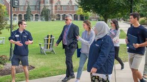 A group of six people walk and talk on a sidewalk near a large brick building with a clock tower and green lawn.