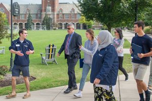 A group of six people walk and talk on a sidewalk near a large brick building with a clock tower and green lawn.