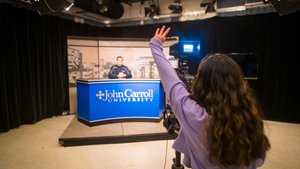 A person with long hair raises their hand while filming another person sitting behind a John Carroll University news desk.