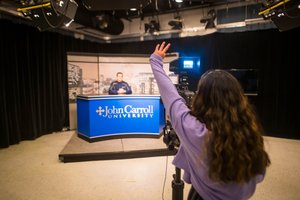 A person with long hair raises their hand while filming another person sitting behind a John Carroll University news desk.