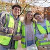 Three young people wearing bright yellow safety vests smile at the camera while standing behind a table with baked goods.