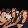 Three young women smile and lean on a barrier at a nighttime outdoor event with a crowd behind them.