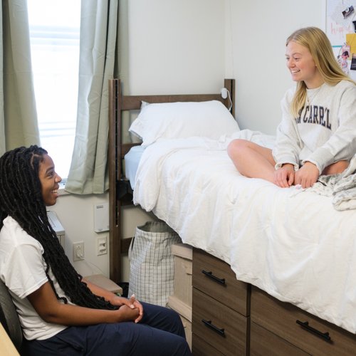Two young women smile and talk in a dorm room, one sitting on a chair and the other on a bed.