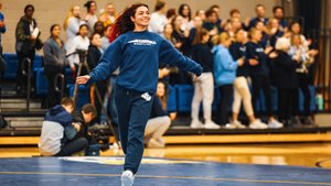A young woman in a blue sweatshirt and sweatpants joyfully runs across a wrestling mat while a crowd watches and claps.