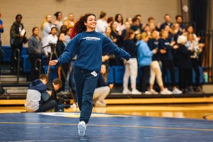 A young woman in a blue sweatshirt and sweatpants joyfully runs across a wrestling mat while a crowd watches and claps.