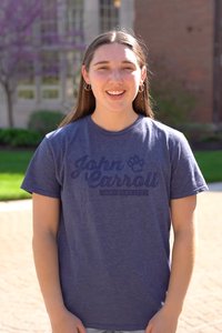A young woman wearing a blue John Carroll University t-shirt and shorts stands smiling outdoors in front of a building.