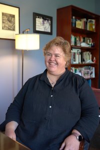 A smiling person with short, light brown hair looks left, seated in a room with a lamp and bookshelf. They appear engaged.