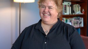 A smiling person with short, light brown hair looks left, seated in a room with a lamp and bookshelf. They appear engaged.