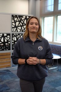 A person stands indoors with hands clasped, wearing a dark pullover with a circular logo and bracelets.