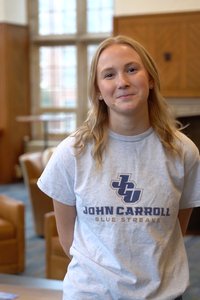 A young woman with blonde hair smiles gently while standing indoors in front of large windows and wooden paneling.