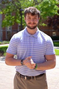 A young man with short brown hair and a beard stands outdoors clasping his hands and smiling.