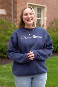 A smiling young woman stands outside in front of a brick building wearing a navy sweatshirt and light jeans.