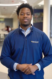 A man wearing a blue John Carroll University pullover stands indoors with his hands clasped in front of him.