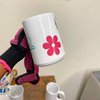 A hand wearing a black glove with pink dots holds a white mug decorated with a pink flower and a blue cartoon character.
