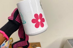 A hand wearing a black glove with pink dots holds a white mug decorated with a pink flower and a blue cartoon character.