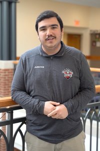 A man with dark hair and a mustache stands indoors, leaning on a railing with his hands clasped.