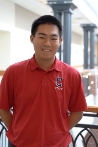 A young man wearing a red polo shirt with a staff logo smiles while standing indoors near a railing.