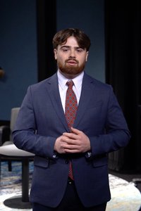 A man with brown hair and beard stands indoors wearing a blue suit and patterned red tie with hands clasped.
