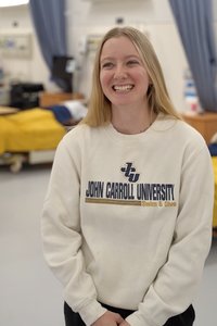 A young woman with long blonde hair smiles while standing in a room with beds covered in yellow blankets.