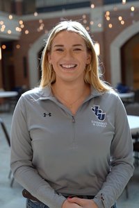 A smiling woman with blonde hair stands indoors wearing a gray zip-up shirt with a swimming and diving logo.